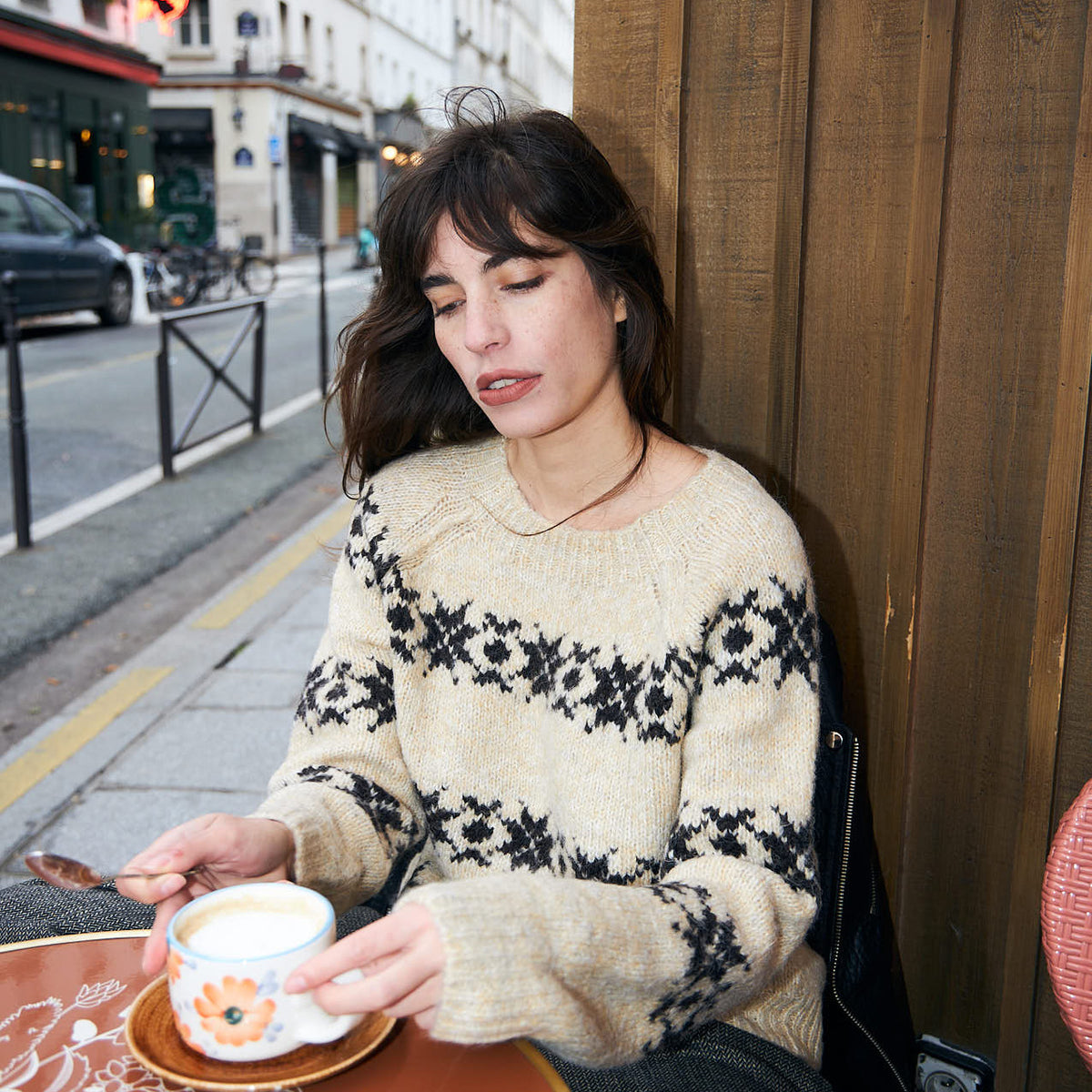 Frau in einem beigen Strickpullover mit nordisch inspiriertem Muster, die in einem Café sitzt und Kaffee trinkt.