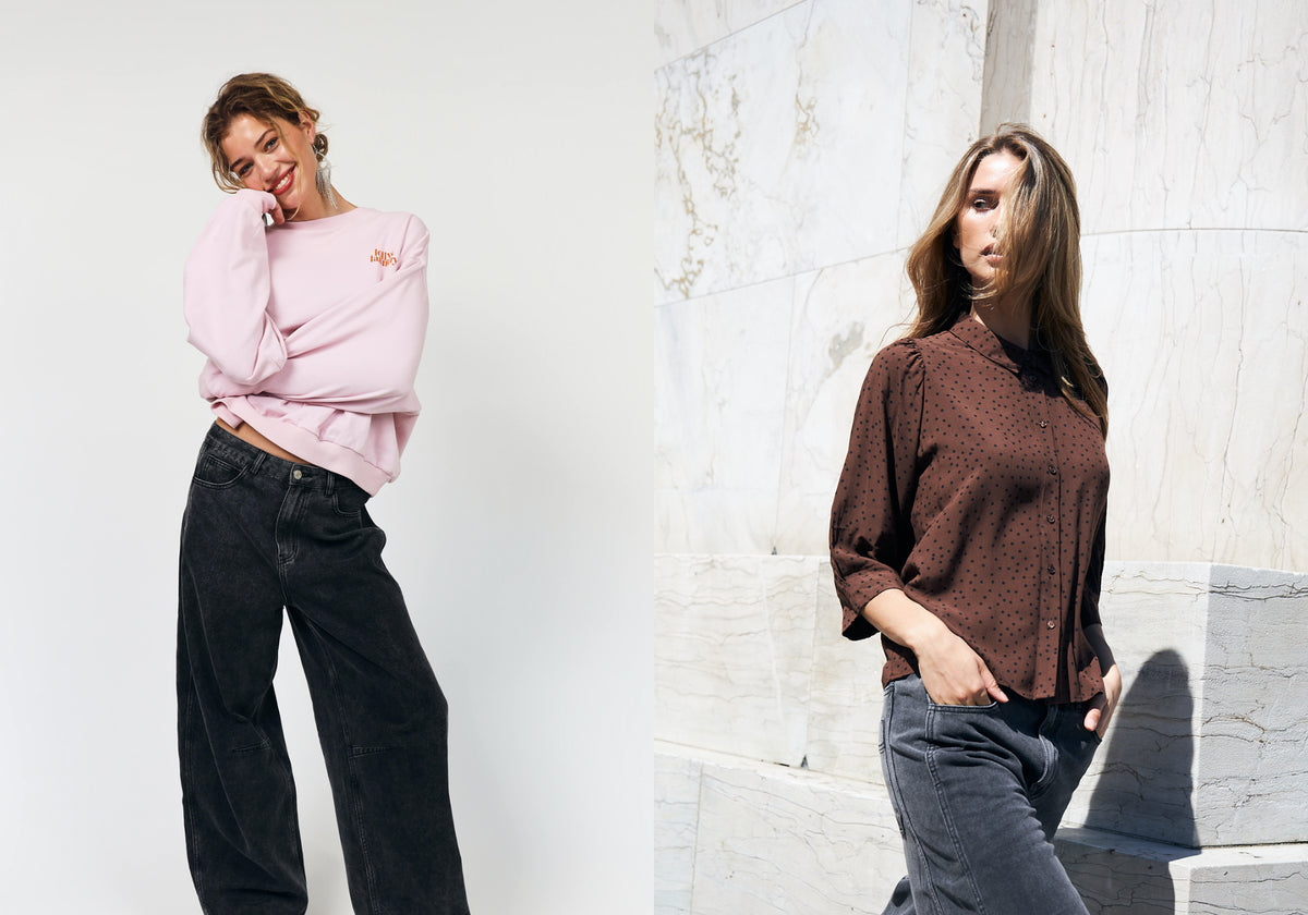 Zwei Frauen in lässigen Outfits. Eine trägt ein hellrosa Sweatshirt mit dunkelgrauen Jeans, während die andere eine braune Bluse mit kleinen schwarzen Punkten und grauen Jeans kombiniert.