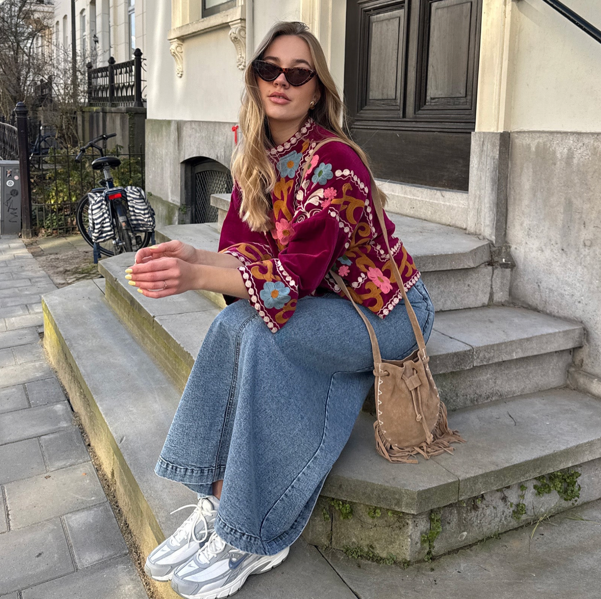 Frau in einer bordeauxroten Jacke von Lollys Laundry mit farbenfrohen Blumenstickereien und dekorativen Borten, kombiniert mit weiter Jeans, Sneakern und brauner Umhängetasche.