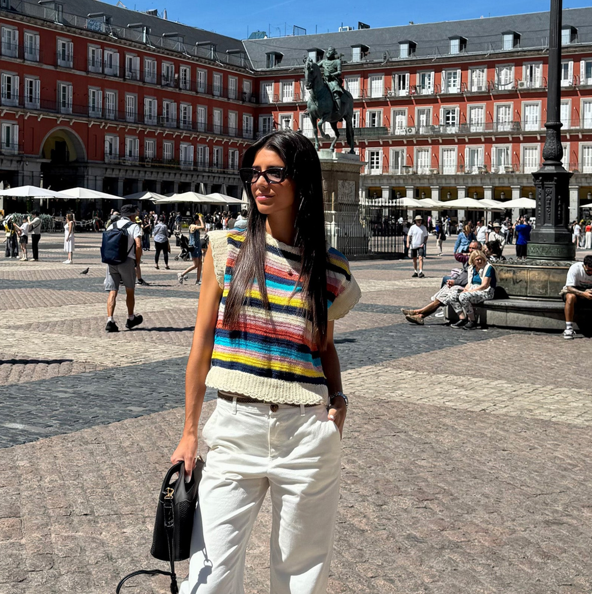 Frau in einem bunten Stricktop mit horizontalen Streifen, kombiniert mit weißer Hose, Sonnenbrille und schwarzer Tasche auf einem sonnigen Stadtplatz.