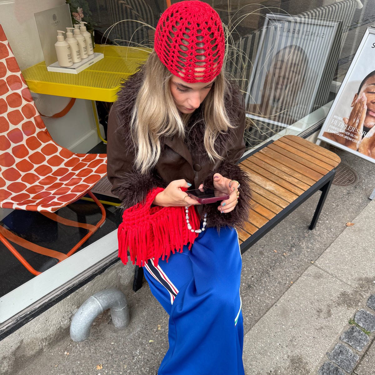 Eine Frau sitzt auf einer Holzbank vor einem Geschäft und blickt auf ihr Handy; sie trägt eine leuchtend rote Häkelmütze, eine braune Kunstfelljacke, eine rote Tasche mit Fransen, einen blauen Rock sowie Schuhe mit Leopardenmuster.