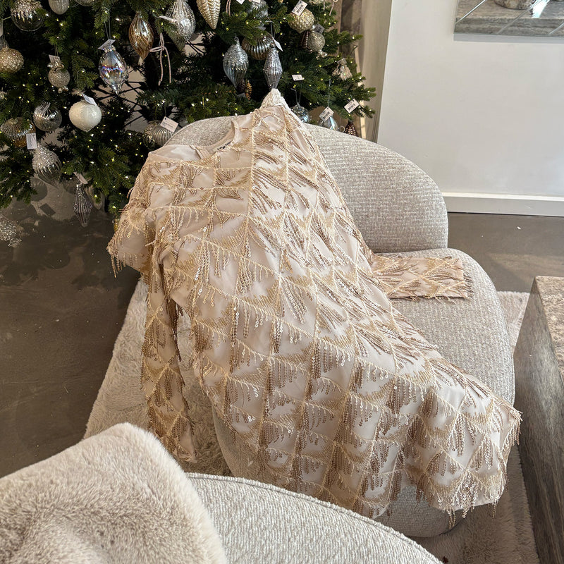 Ein champagnerfarbiges Kleid mit goldfarbenen Perlendetails liegt über einem hellgrauen Loungesessel drapiert, vor einem geschmückten Weihnachtsbaum mit silbernen und gläsernen Ornamenten.