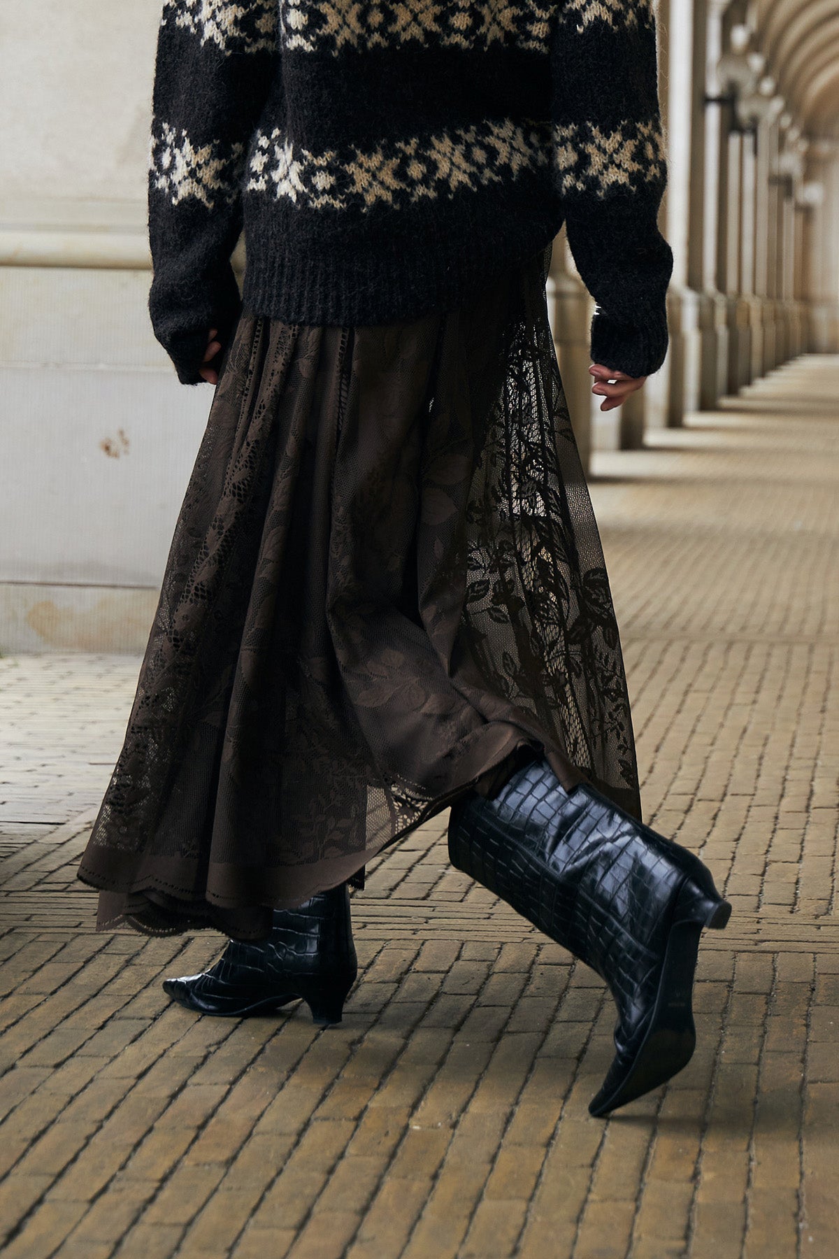 Frau in einem braunen Strickpullover mit nordisch inspiriertem Muster, kombiniert mit einem braunen Spitzenrock und schwarzen Stiefeln in Kopenhagen.