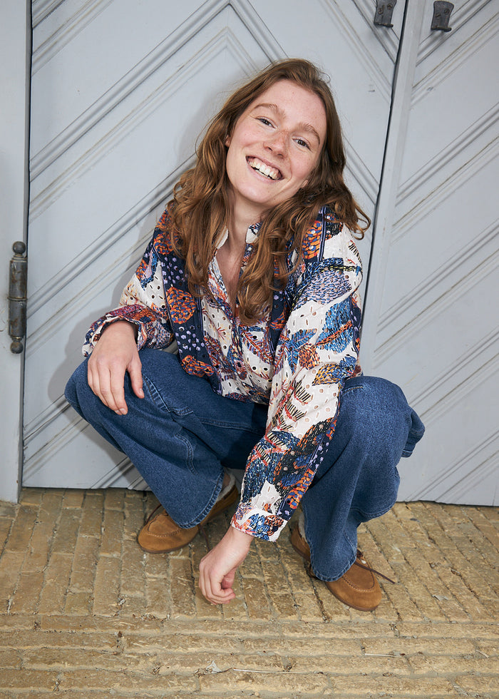 Frau in farbenfrohem Hemd mit Blattmuster und Lochdetails in Blau, Creme und Orange, kombiniert mit blauen Jeans und braunen Schuhen.