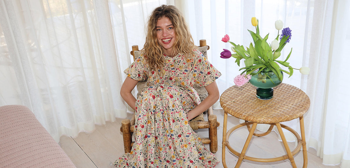 Lächelnde Frau in einer Bluse und einem passenden Rock mit buntem Blumenmuster, die auf einem Holzstuhl neben einem Rattantisch mit Tulpen sitzt.