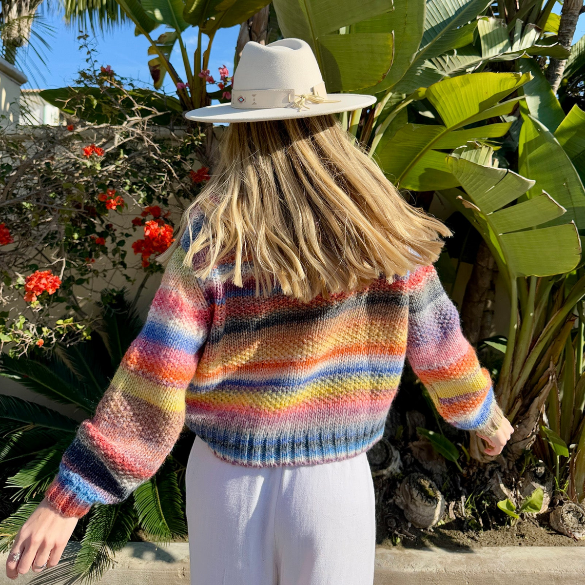 Frau im farbenfrohen FairhavenLL Strickpullover von Lollys Laundry mit bunten Streifen in Blau, Pink, Orange und Gelb, kombiniert mit heller Hose und beigem Hut.