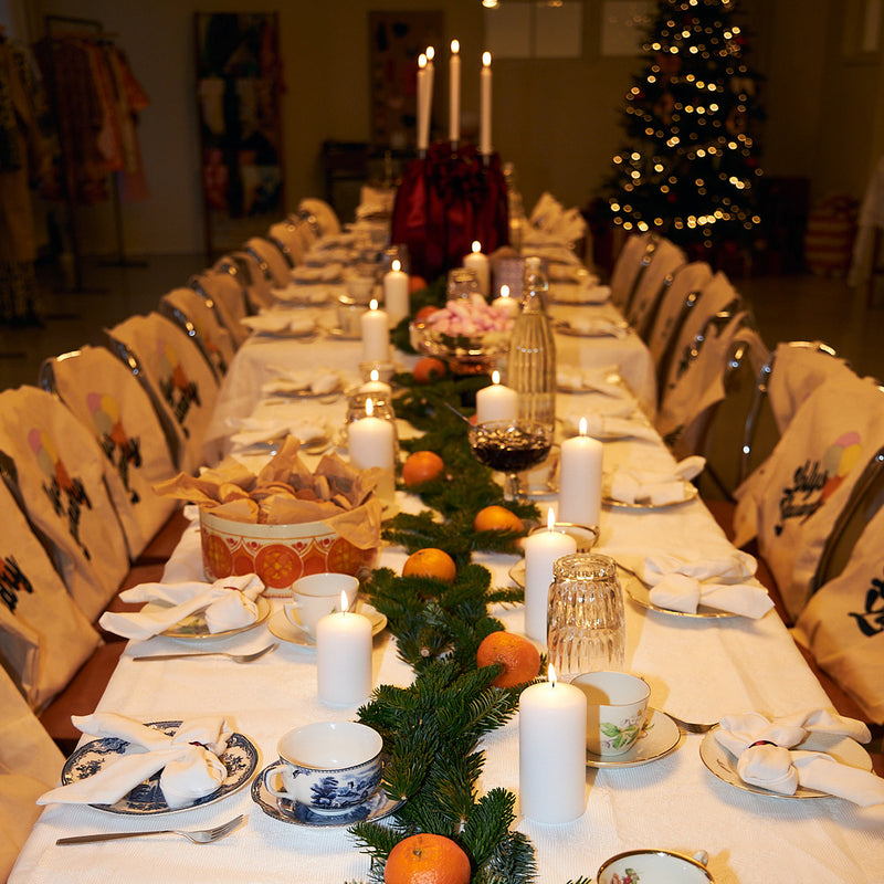 Weihnachtlich gedeckte lange Tafel mit brennenden Kerzen, Tannenzweigen, Orangen und gemustertem Porzellan. Im Hintergrund leuchtet ein Weihnachtsbaum.