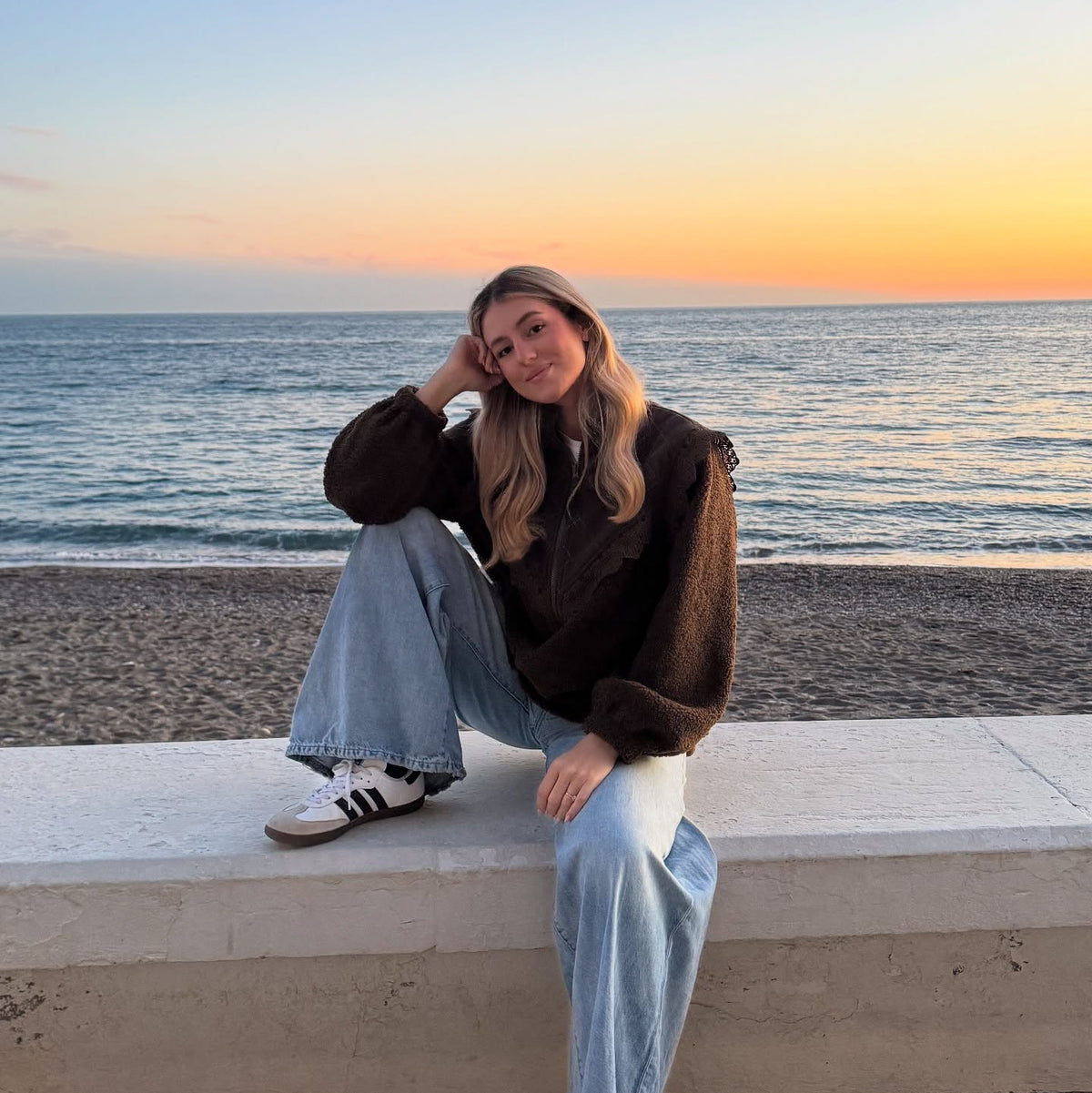 Frau in einer dunkelbraunen Teddyjacke mit weicher Struktur, hellblauer Jeans und Sneakern, die bei Sonnenuntergang am Strand sitzt.