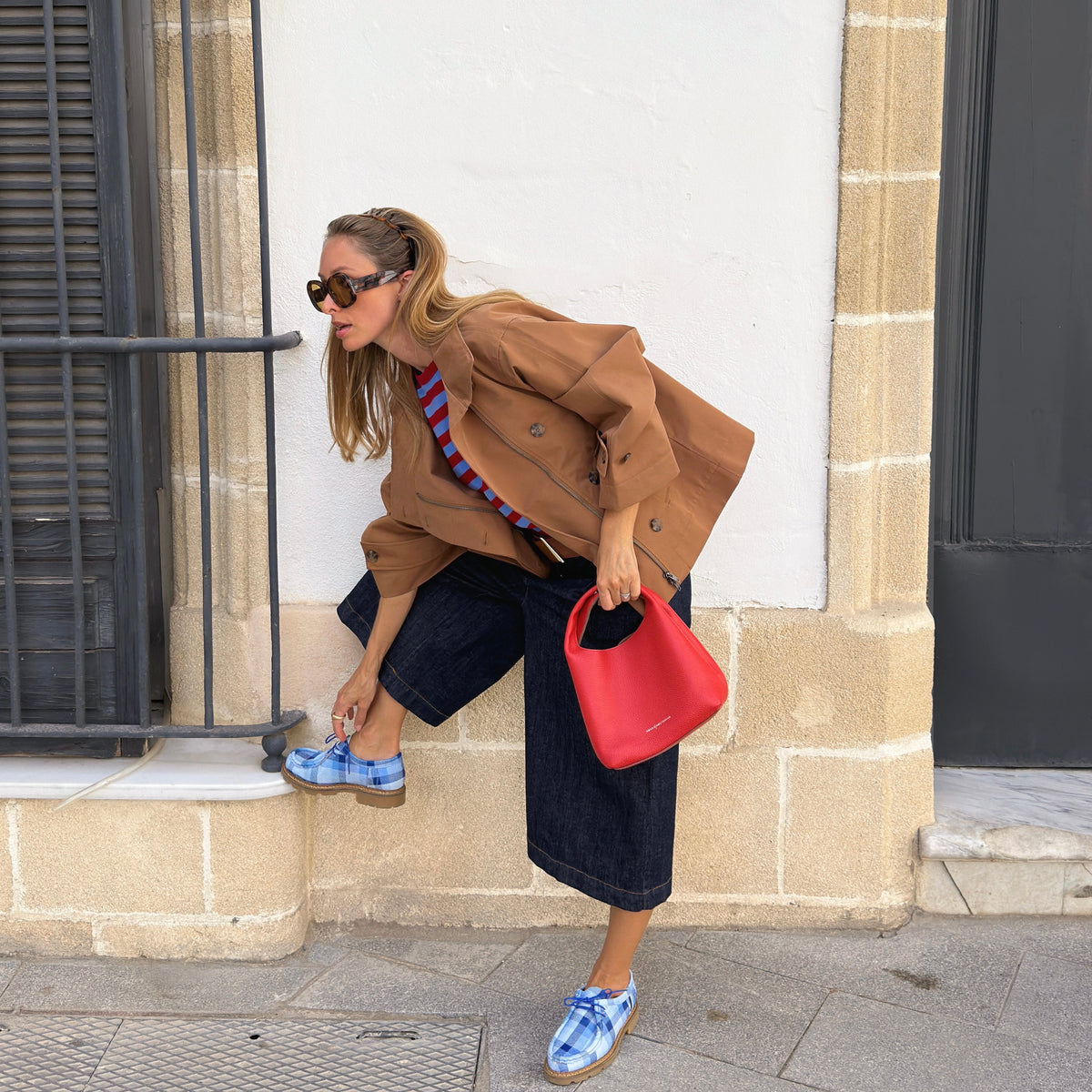 Frau in einer braunen Jacke, einem rot-blau gestreiften Oberteil und dunklen Denim-Culottes, kombiniert mit blau karierten Schuhen, einer Sonnenbrille und einer roten Tasche.