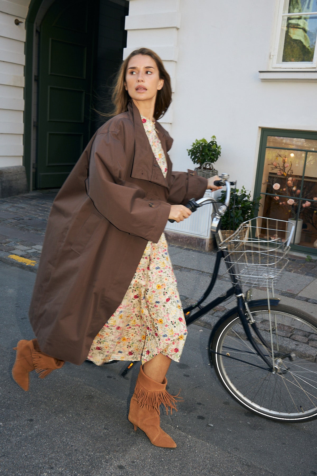 Frau mit einem Fahrrad auf einer Straße in Kopenhagen. Sie trägt einen langen braunen Mantel über einem geblümten Kleid und hohe Stiefel mit Absatz.