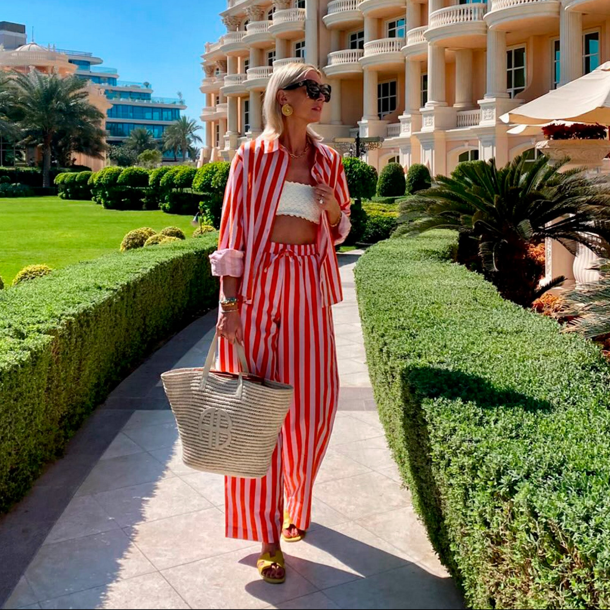 Frau in einem rot-weiß gestreiften Set von Lollys Laundry mit Hemd und passender Hose, kombiniert mit weißem Top, Sonnenbrille und geflochtener Tasche.