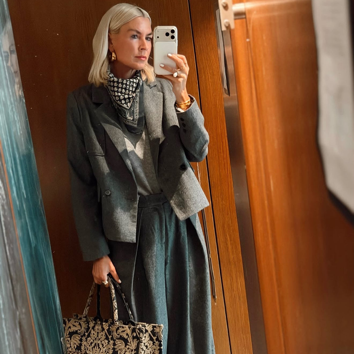 Eine Frau macht ein Spiegel-Selfie in einem Innenraum mit holzvertäfelten Wänden; sie trägt einen grauen Blazer, einen gemusterten Schal sowie eine graue Hose mit weitem Bein und hält eine schwarz-beige gemusterte Tragetasche.