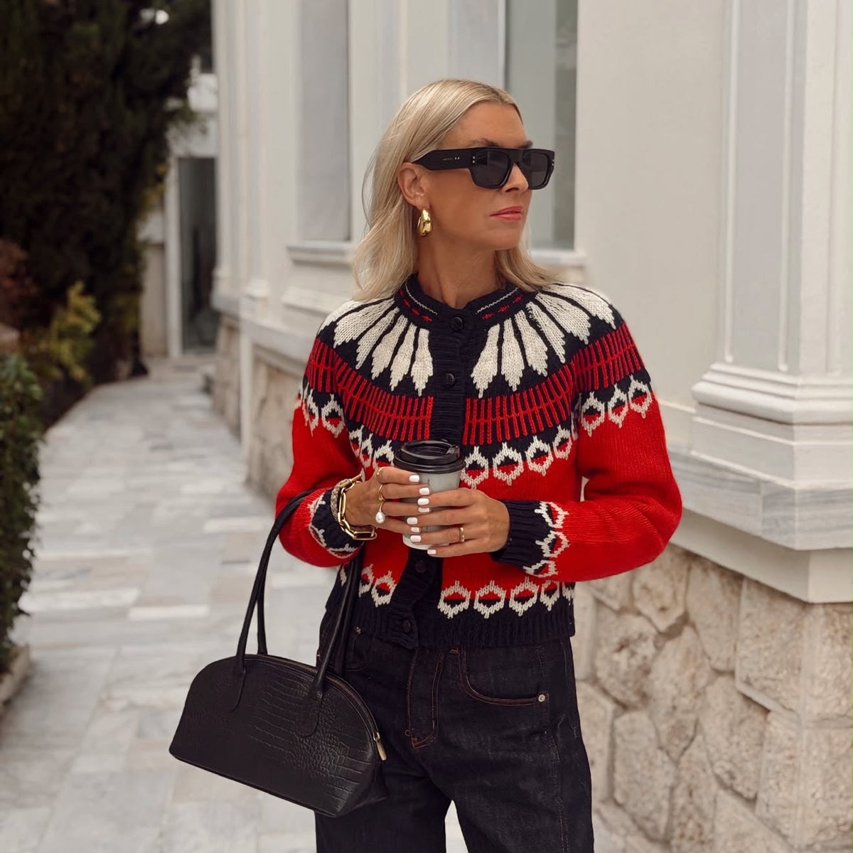 Frau in der ClareLL Strickjacke von Lollys Laundry mit rot-schwarzem und cremefarbenem Muster, kombiniert mit dunkler Jeans, Sonnenbrille und schwarzer Handtasche.