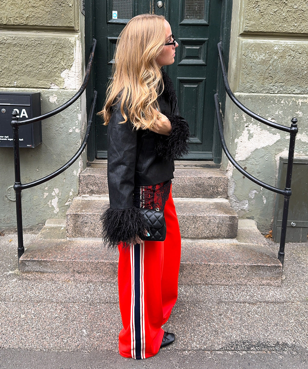 Frau in einer schwarzen Jacke mit flauschigen Ärmeldetails und einer roten Hose mit seitlichen Streifen, kombiniert mit Sonnenbrille und schwarzer Tasche vor einer grünen Tür.