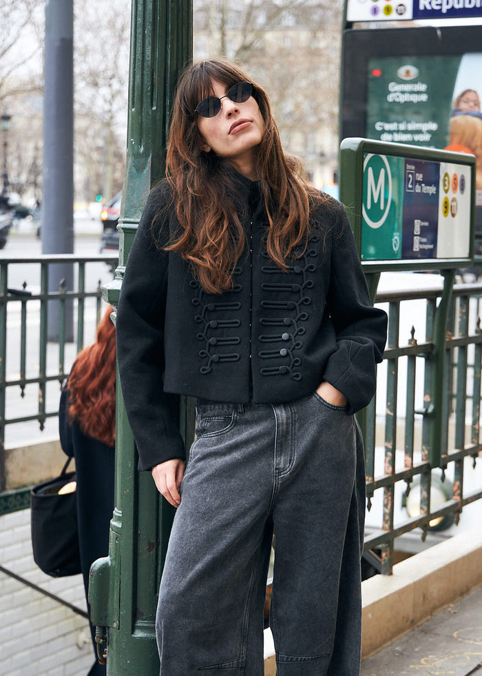 Frau in kurzer schwarzer Napoleonjacke mit dekorativen Details, kombiniert mit dunkelgrauen Jeans und schwarzer Sonnenbrille.