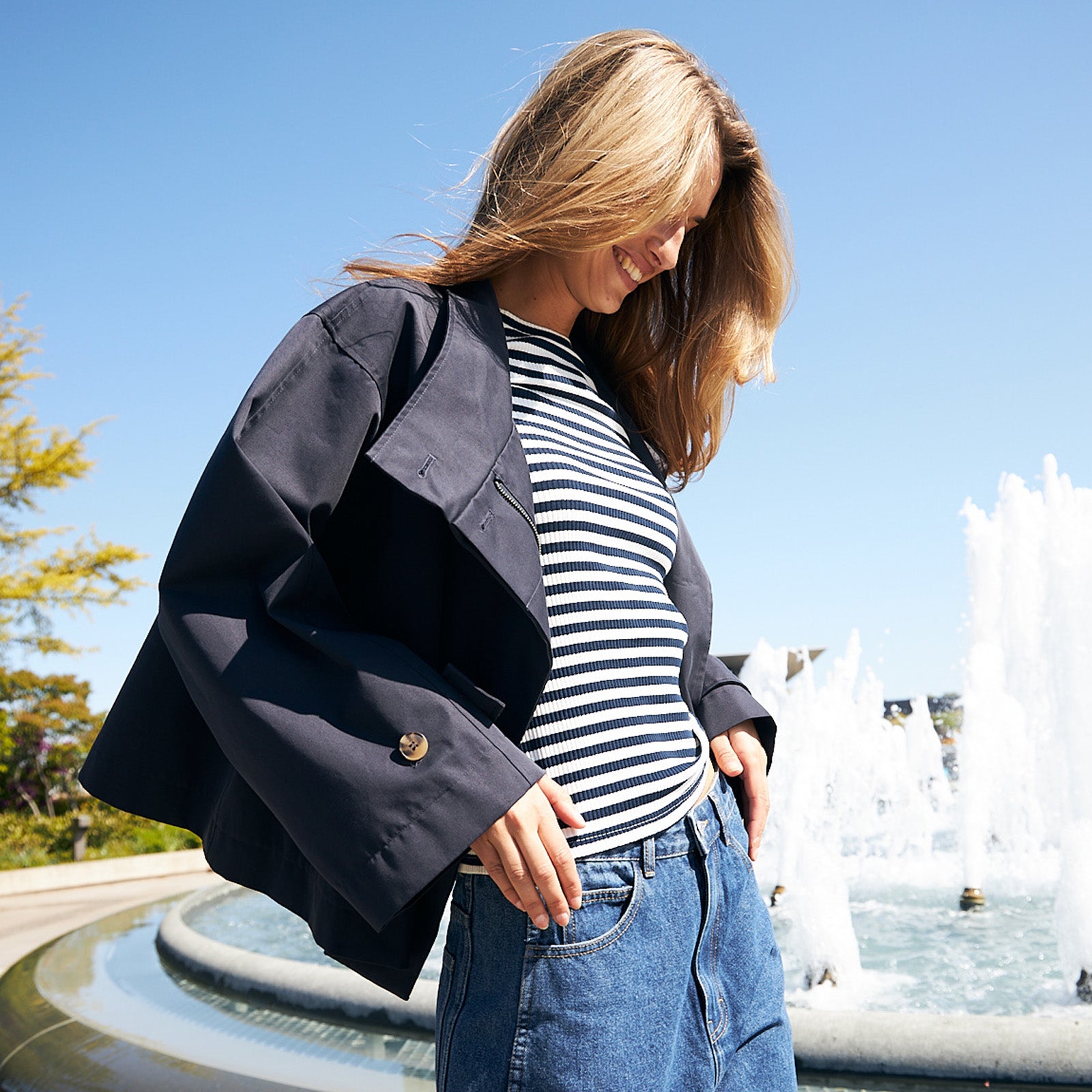 Frau trägt eine dunkelblaue Jacke, ein gestreiftes T-Shirt und eine blaue Jeans und steht an einem Brunnen an einem sonnigen Tag.