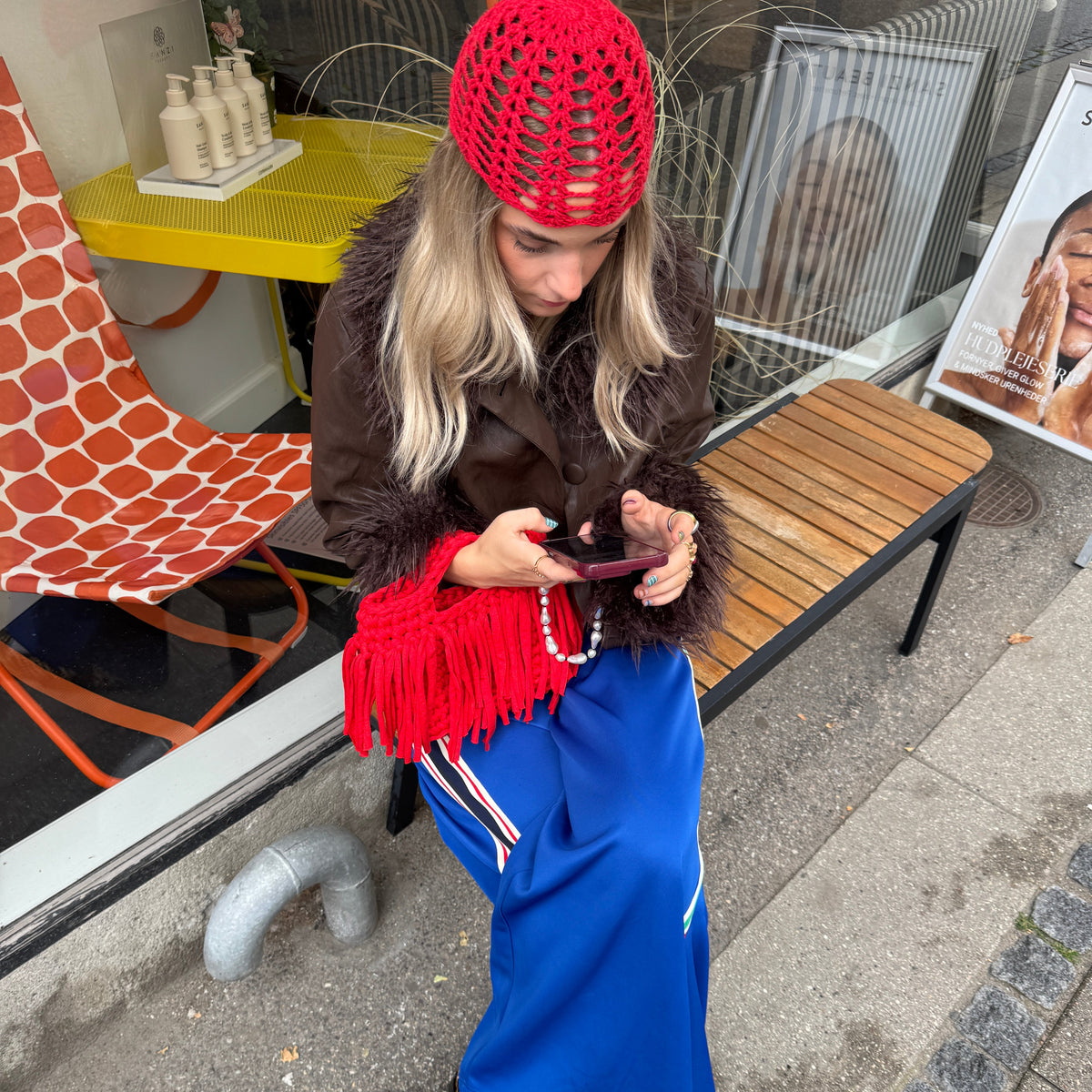 Frau auf einer Bank mit einer braunen Jacke mit Pelzbesatz, einer blauen Hose mit Streifendetails und einer roten Häkelmütze sowie einer roten Fransentasche auf dem Schoß.