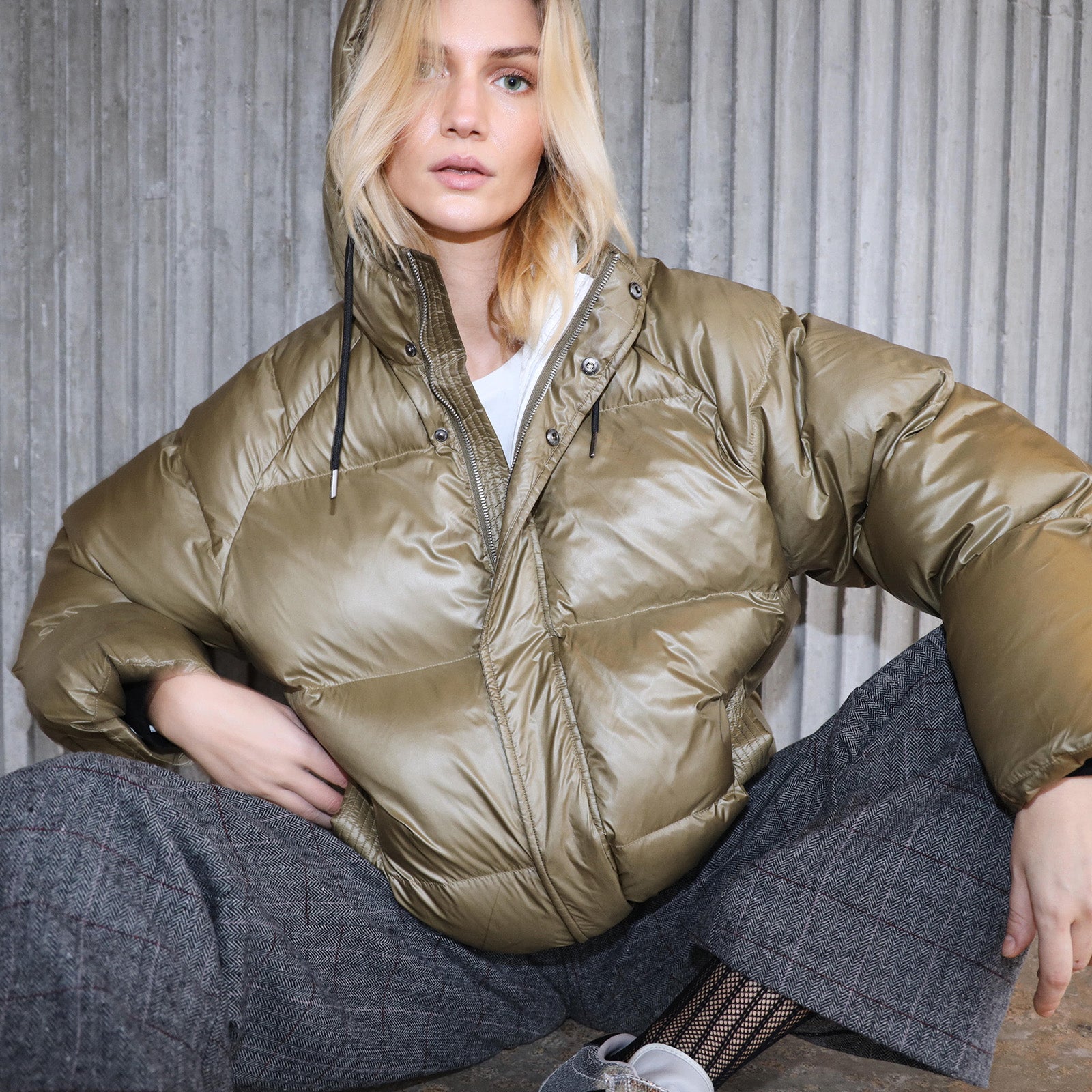 Model trägt eine olivgrüne Steppjacke, kombiniert mit einer karierten grauen Hose vor einem grauen Beton Hintergrund.