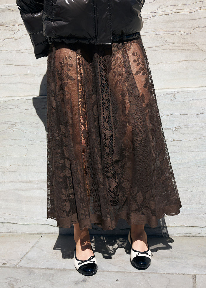 Frau in langem braunem Spitzenrock mit Blumenmuster, kombiniert mit schwarzer Jacke und schwarz weißen Ballerinas.