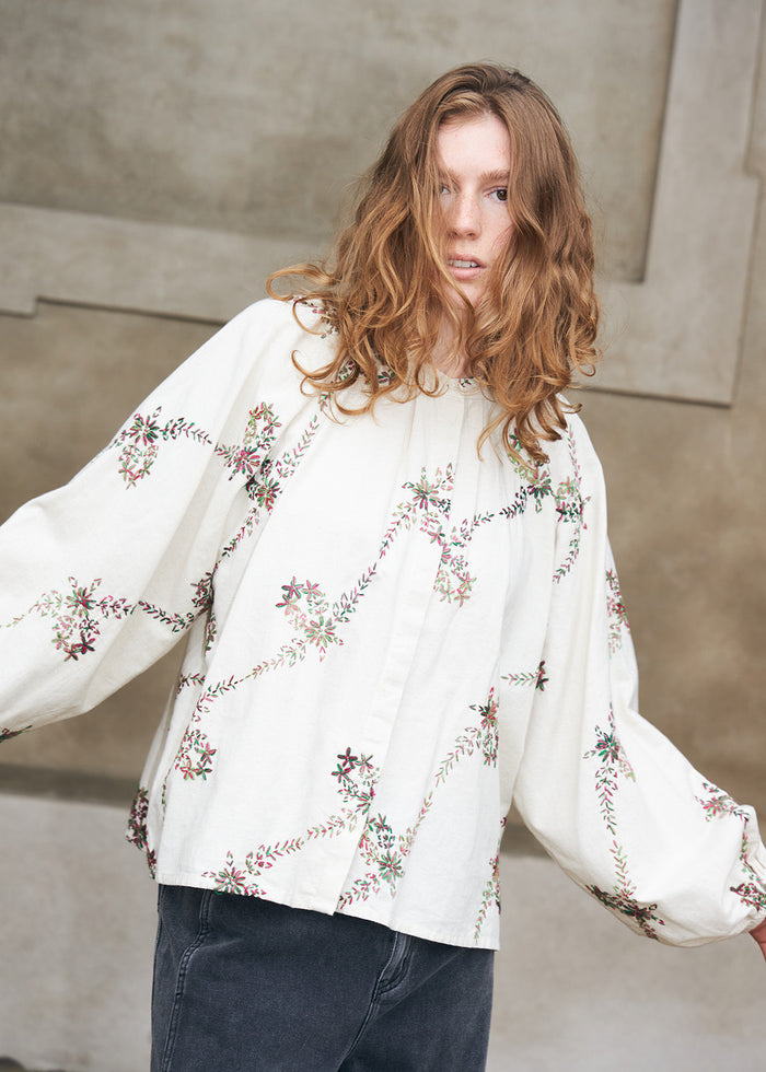 Frau trägt eine weiße Bluse mit farbenfrohen Blumenstickereien und voluminösen Ärmeln, kombiniert mit dunklen Denimjeans. Ein femininer Alltagslook mit dekorativen Details.