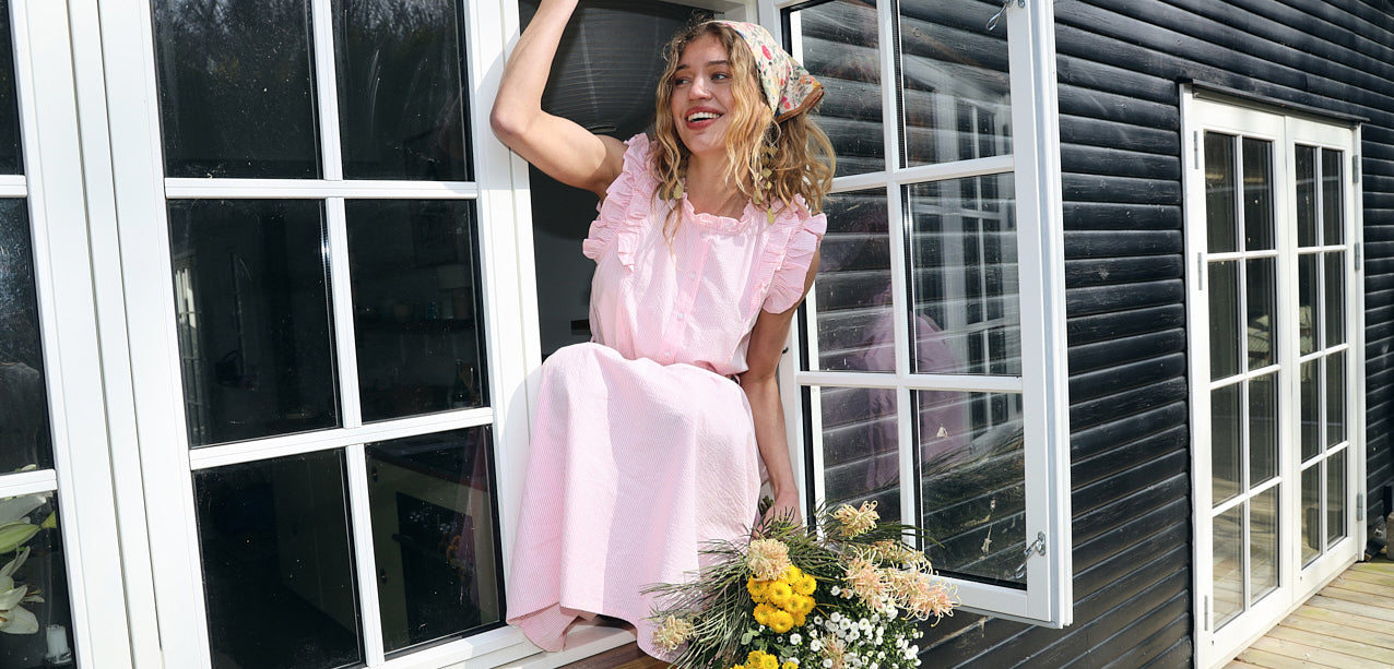 Frau in einem rosafarbenen Kleid mit Rüschendetails und floralem Tuch, sitzend in einem offenen Fenster mit einem Blumenstrauß darunter.