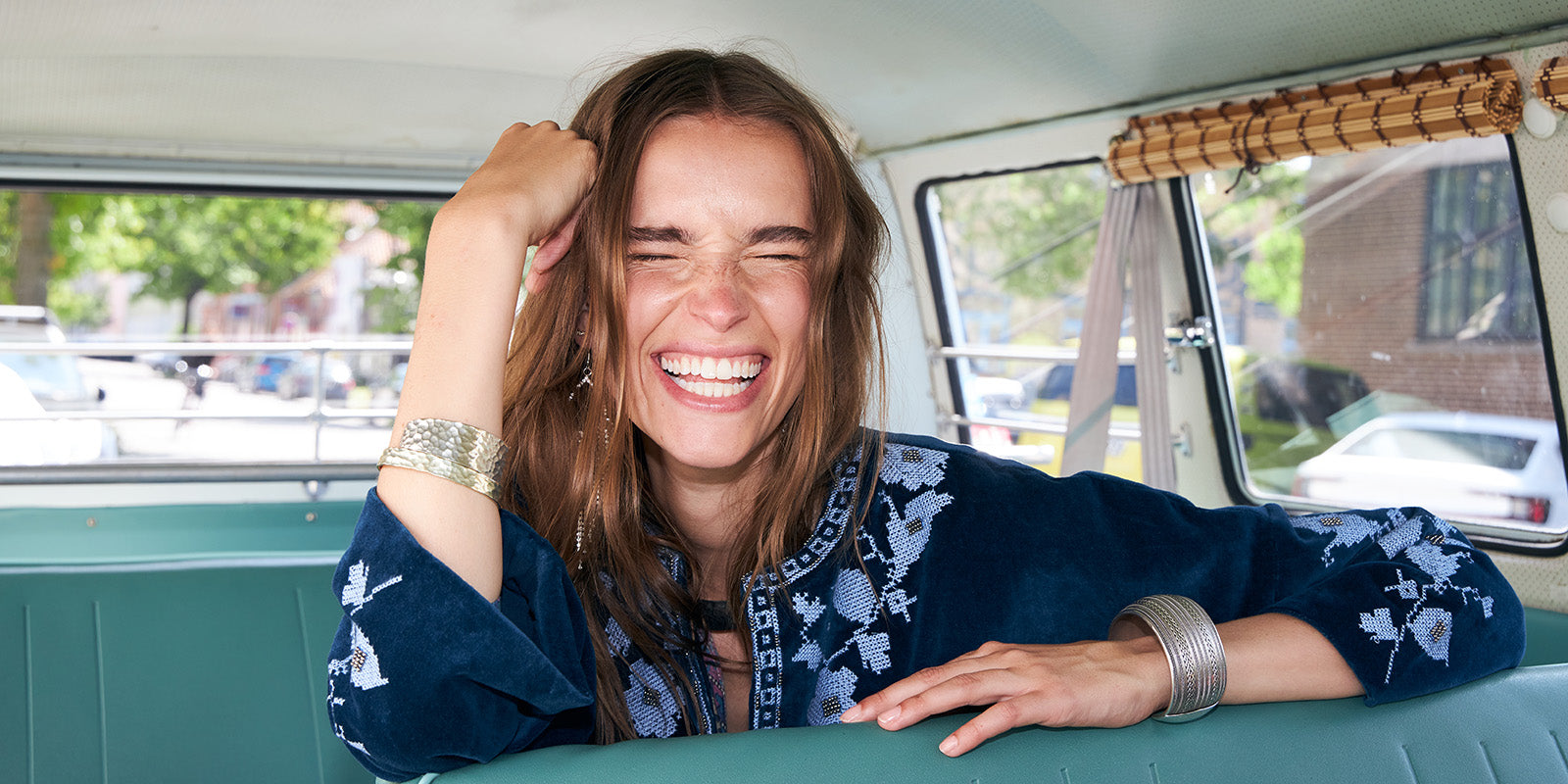 Lächelnde Frau in einem Vintage Camper, gekleidet in einer dunkelblauen Jacke mit hellen Stickereidetails. Das Bild vermittelt sommerliche Leichtigkeit, entspannte Eleganz und einen Boho inspirierten Stil.