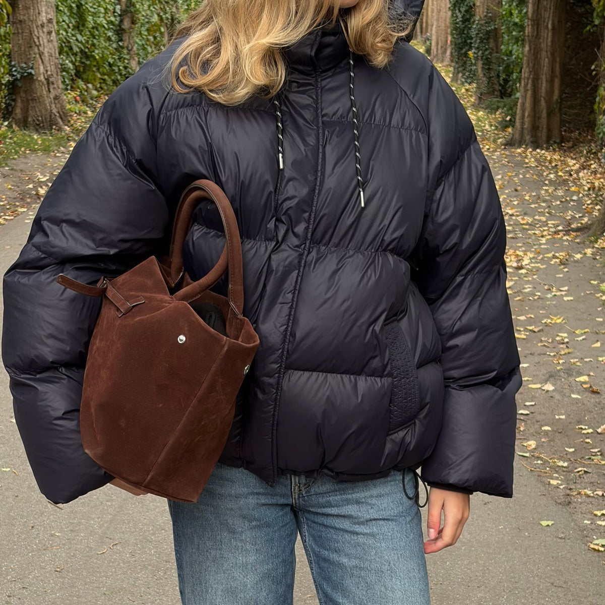 Eine Frau steht im Freien auf einem gepflasterten Weg; sie trägt eine schwarze Daunenjacke, eine blaue Jeans und braune Schuhe und hat eine braune Wildleder-Tragetasche bei sich.