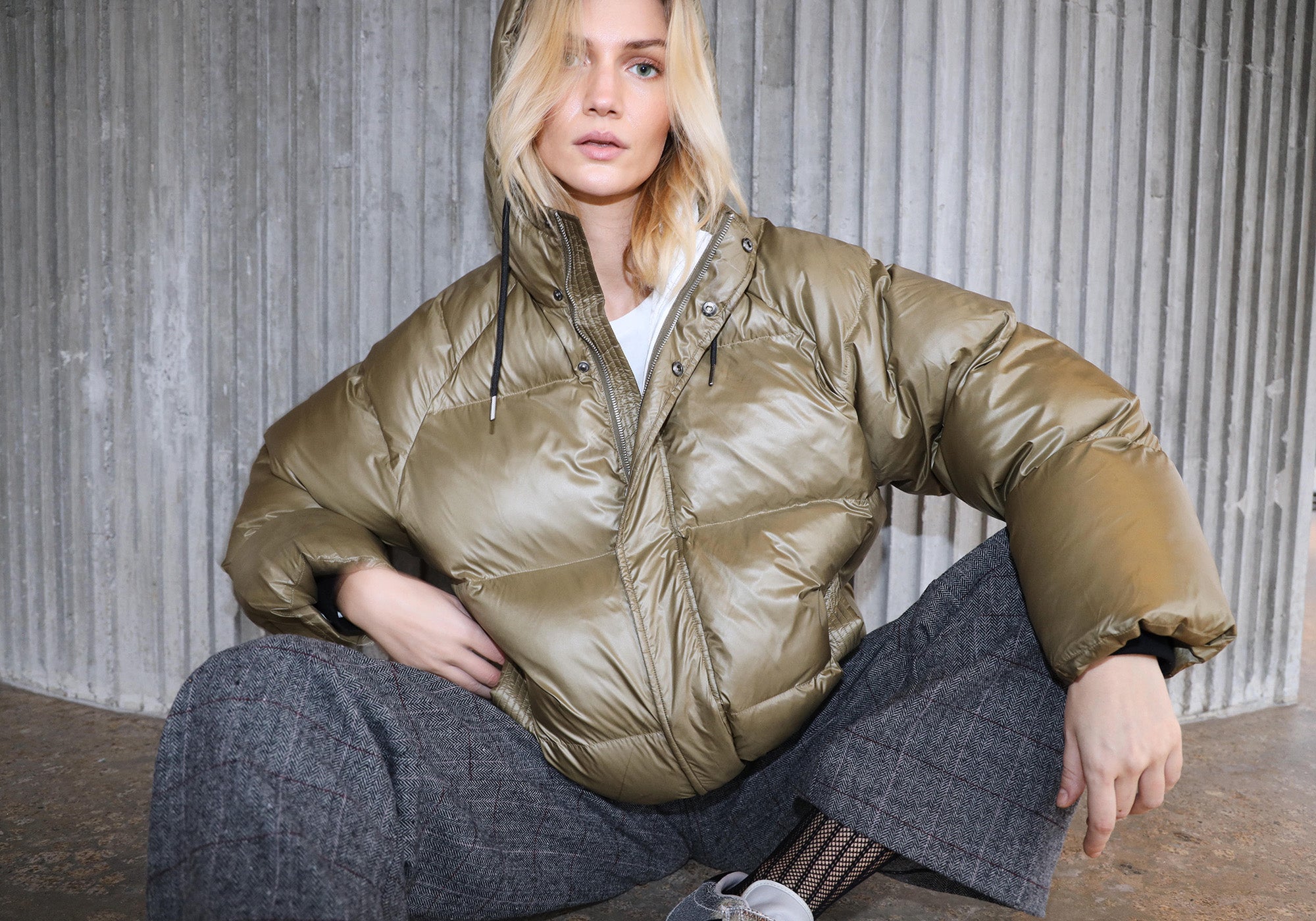 Model trägt eine olivgrüne Daunenjacke mit Kapuze, kombiniert mit grauen karrierten Hosen und einem weißen T-Shirt von Lollys Laundry.