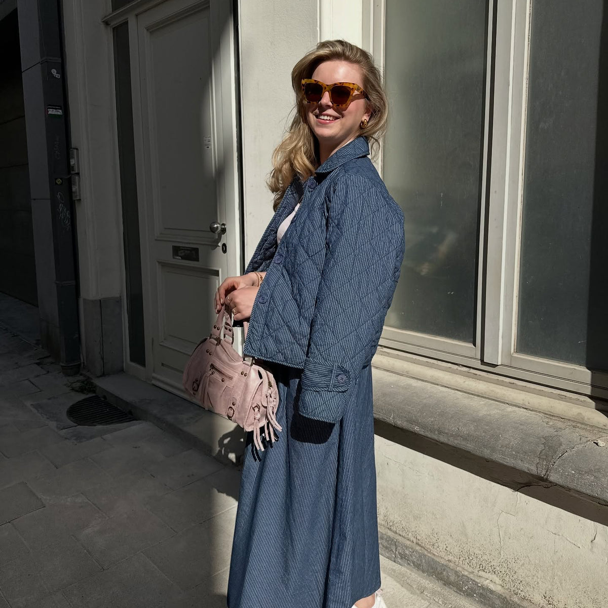 Frau in einer blau gemusterten Jacke mit schwarzen Kontrastdetails, kombiniert mit passender Hose, Sonnenbrille und rosafarbener Handtasche.