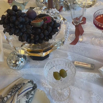 Festlich gedeckter Weihnachtstisch mit Weintrauben, Obst, Kristallgläsern, Discokugeln und dekorativen Details auf einer weißen Tischdecke.