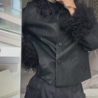 Video einer Frau in einer schwarzen Jacke mit lockigem Fellimitat an Kragen und Ärmelabschlüssen, kombiniert mit einer dunklen karierten Hose.