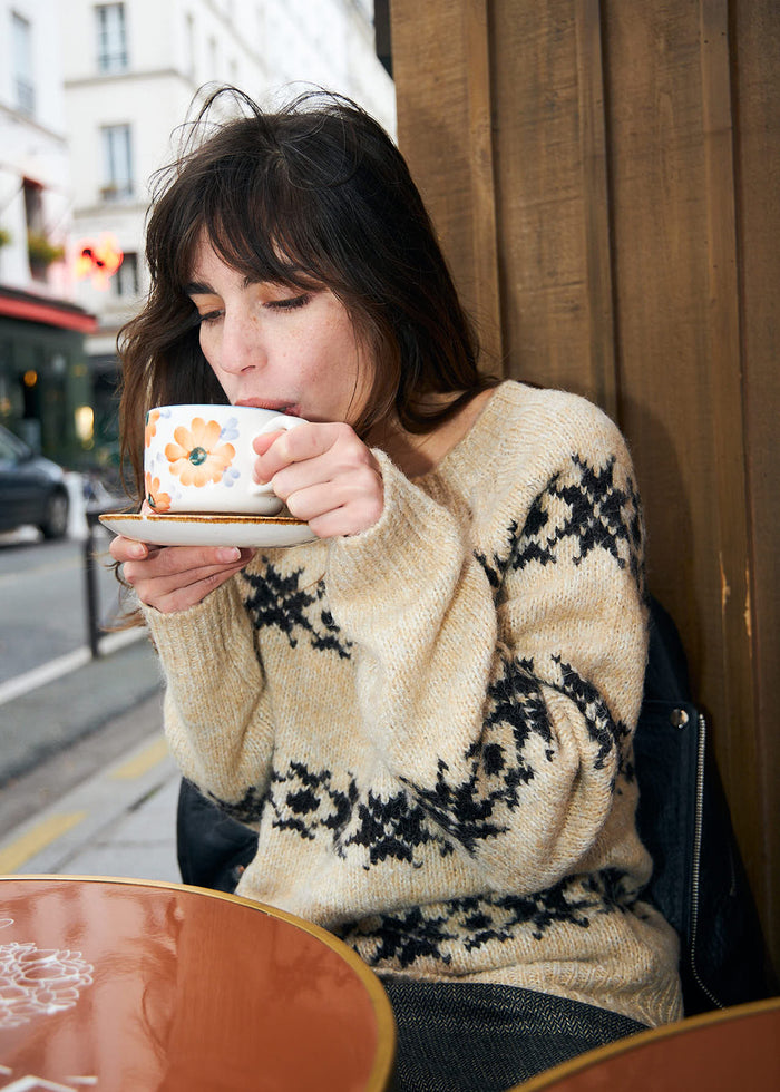 Frau in cremefarbenem Strickpullover mit dunklem nordisch inspiriertem Muster, während sie eine Tasse Kaffee in einem Café genießt.