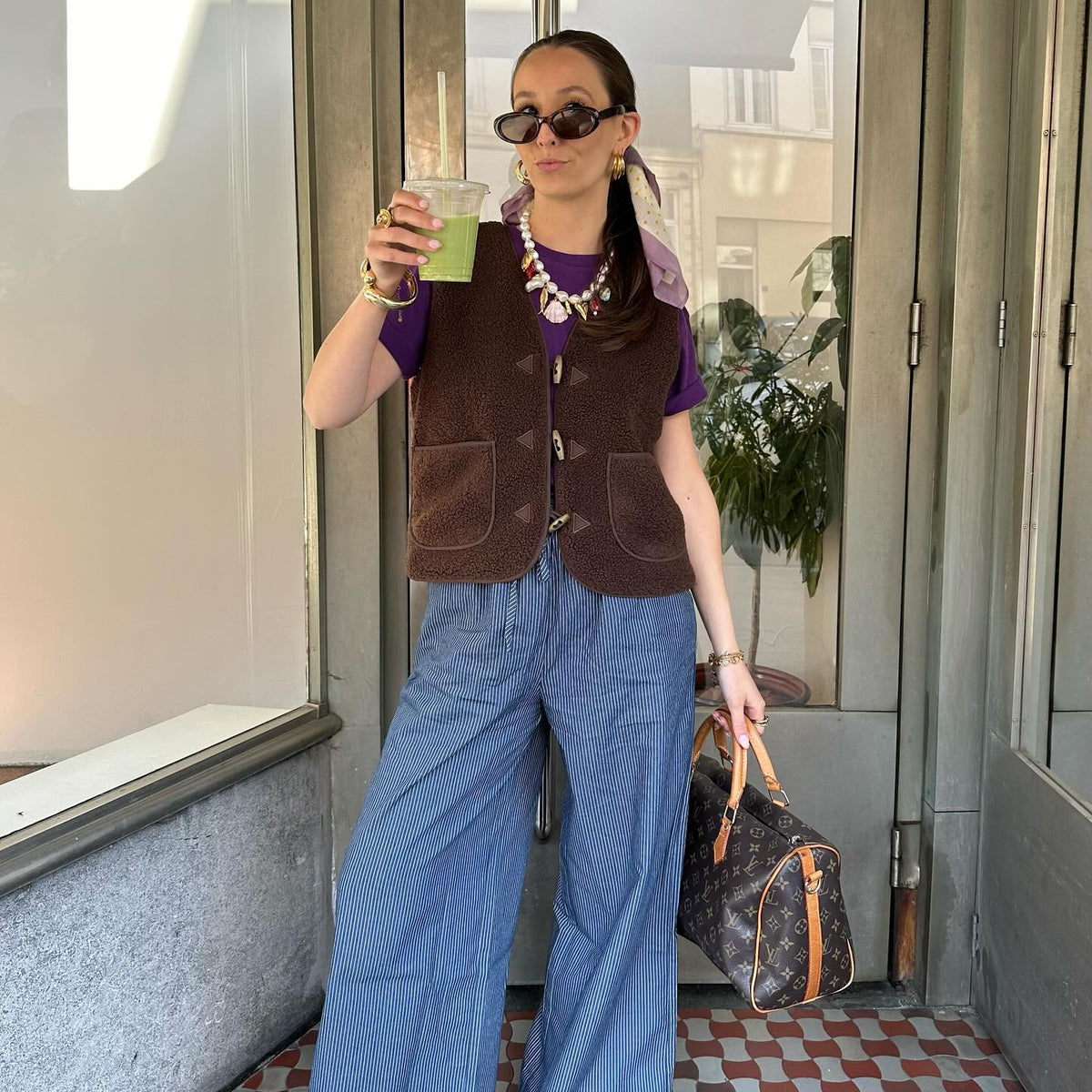 Frau in einer braunen Weste über einem lila T-Shirt und einer weit geschnittenen blau gestreiften Hose, kombiniert mit Sonnenbrille, Schmuck und gemusterter Tasche.